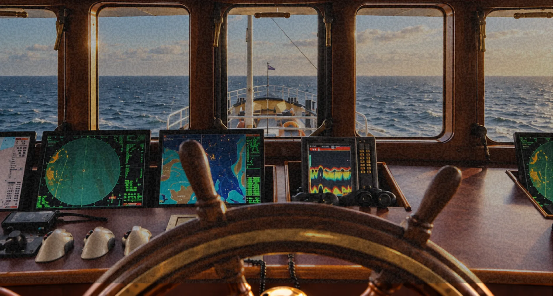 Ship bridge with navigation screens and a wooden helm looking out to sea at sunset