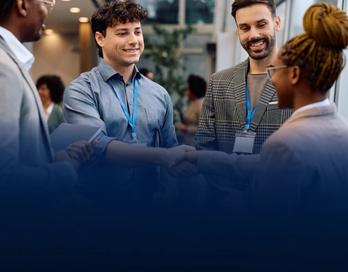 Four diverse professionals wearing lanyards smiling and shaking hands in an office lobby