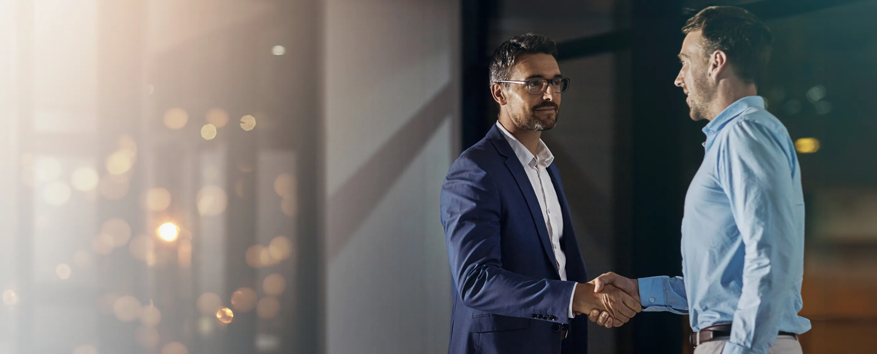 Two businessmen shaking hands in a modern office at night with warm city lights behind them