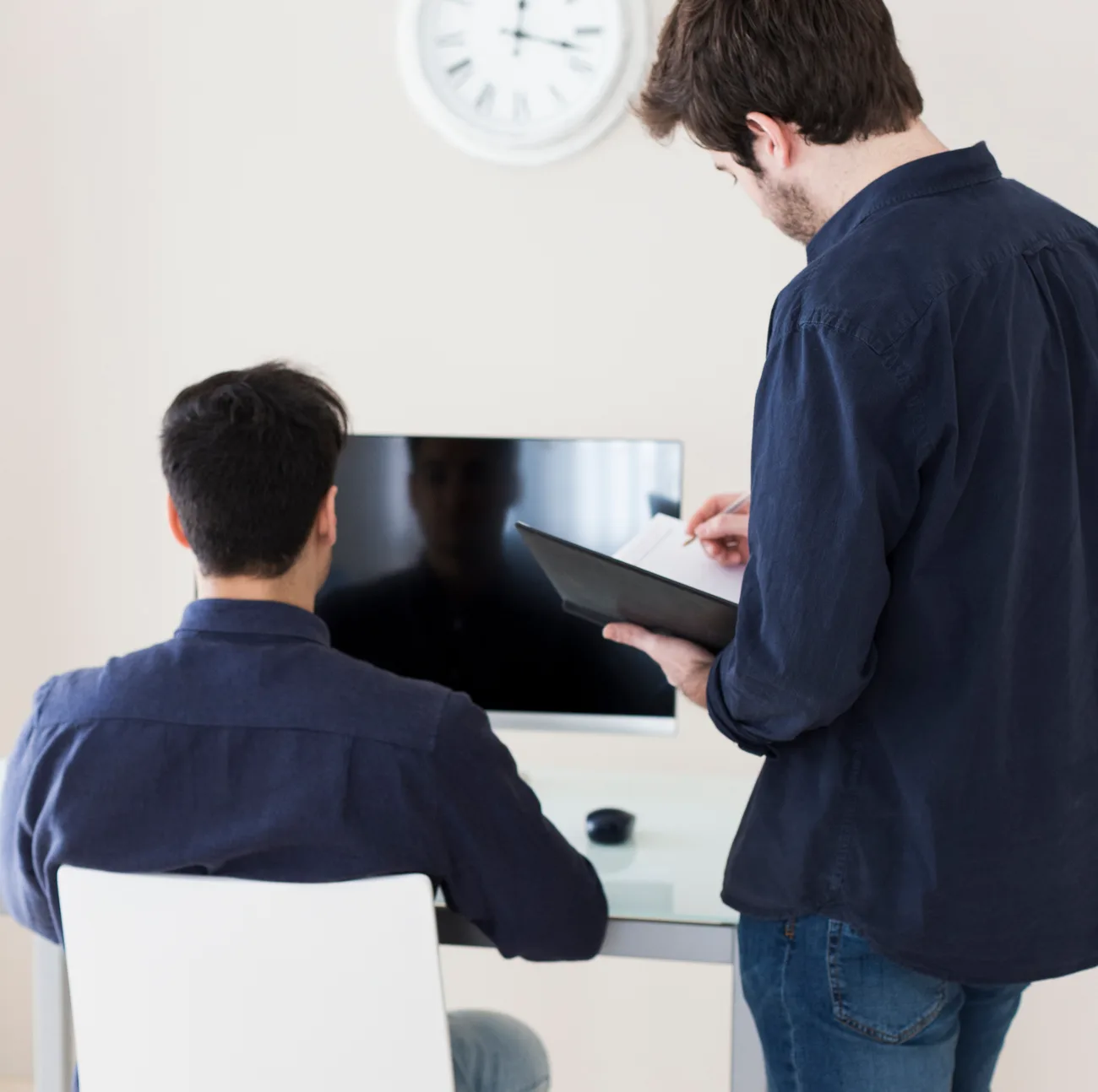 Colleague with a clipboard standing beside a man working at a computer monitor under a wall clock