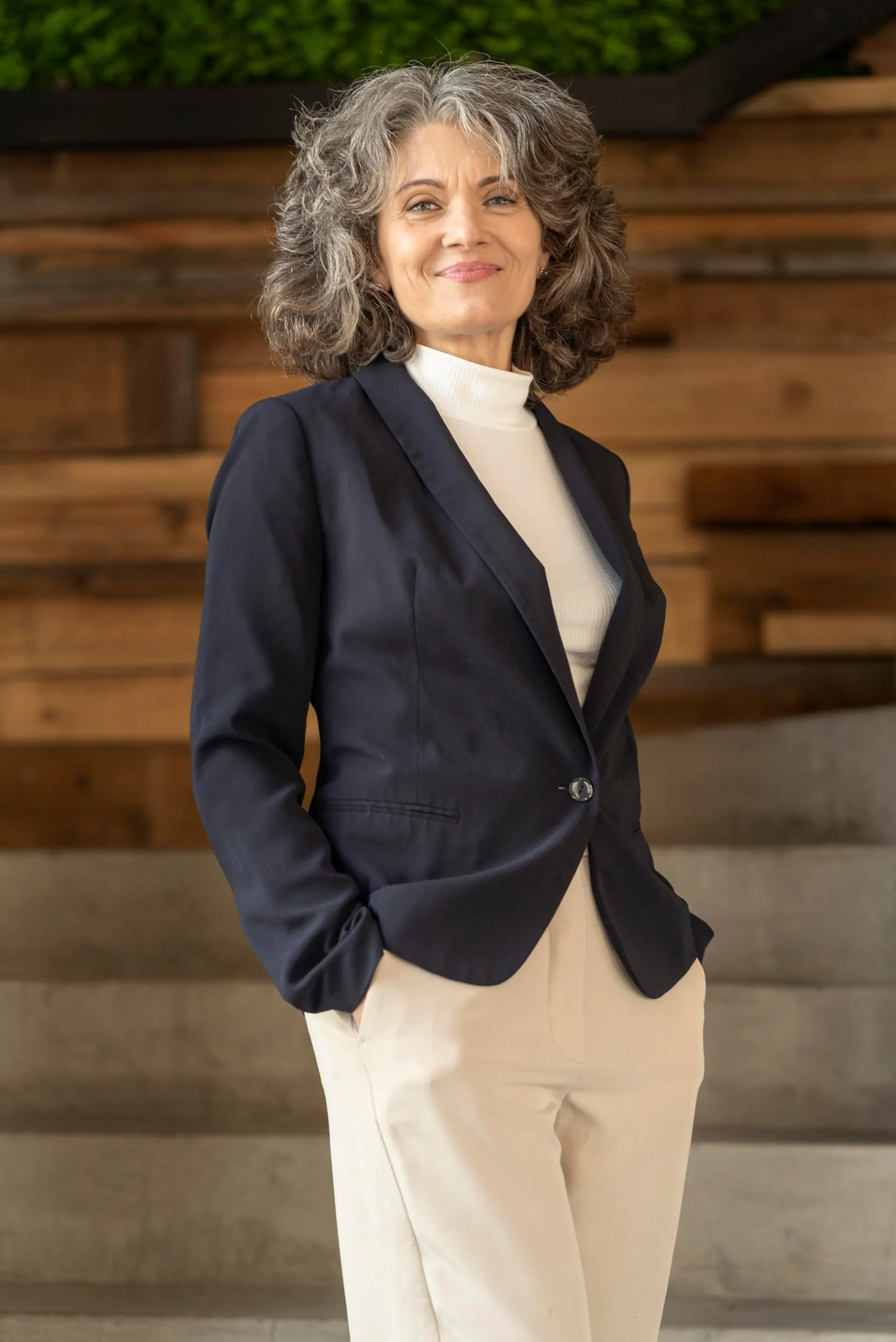 Smiling woman with curly grey hair in a navy blazer and white turtleneck in front of a wooden wall