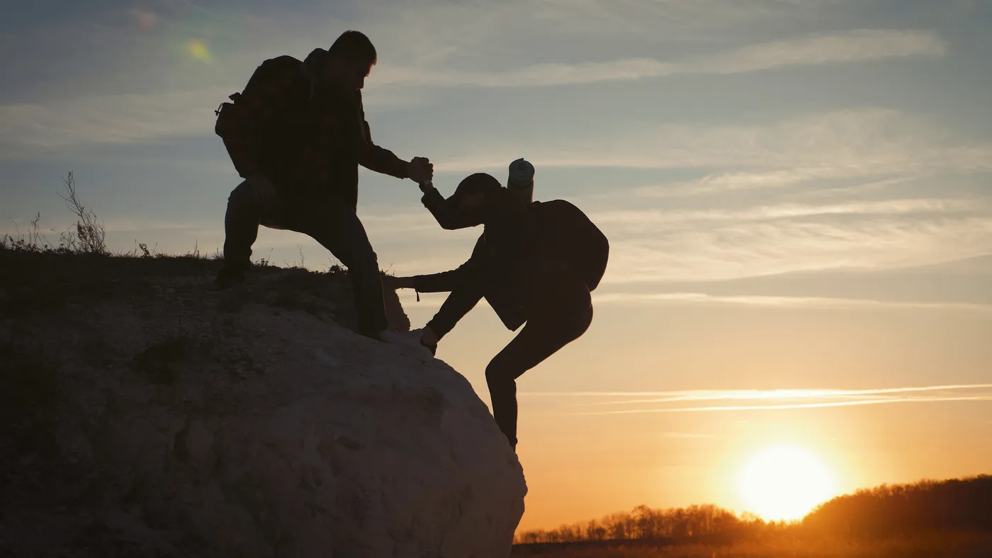 Silhouette of a hiker helping another climb onto a rock at sunset