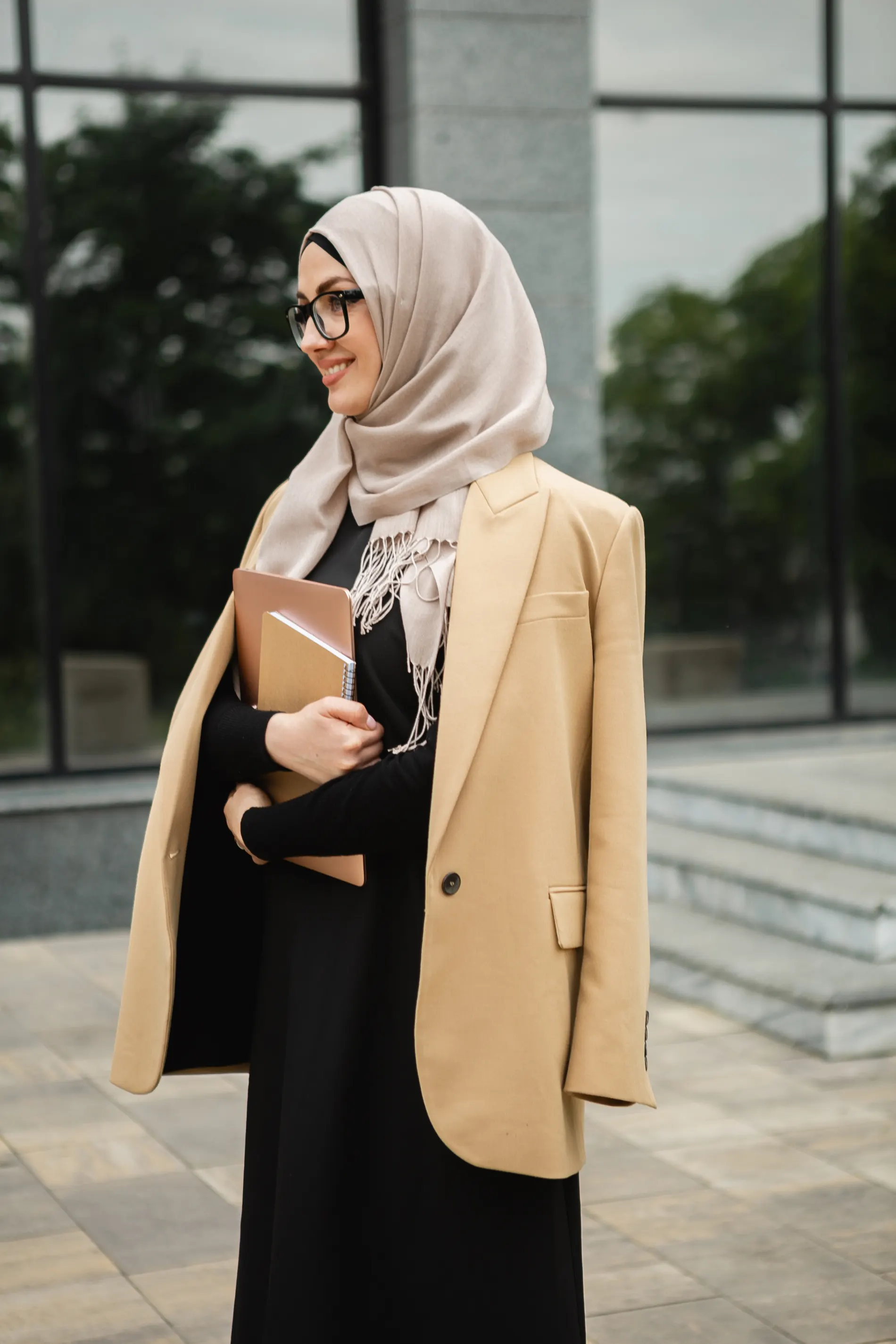 Smiling woman in a beige hijab, glasses and camel blazer holding a notebook outside a glass office building