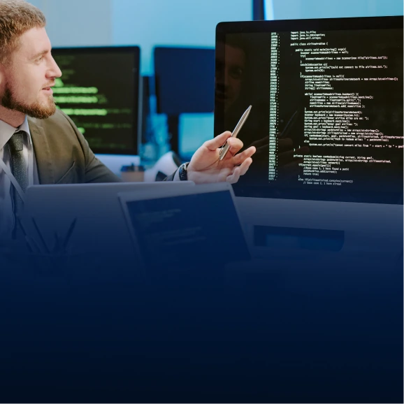 Male engineer sitting at a desk reviewing green code on a large dark monitor