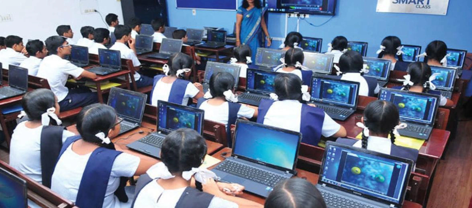 Students in uniform working on laptops in a smart-class computer lab while a teacher presents