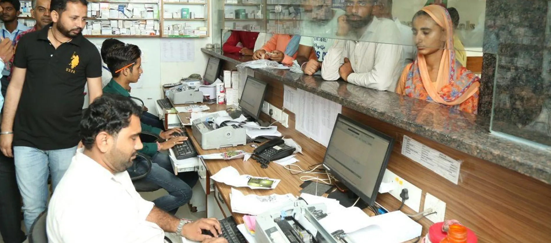 Hospital billing counter with staff at computers and patients waiting behind the glass