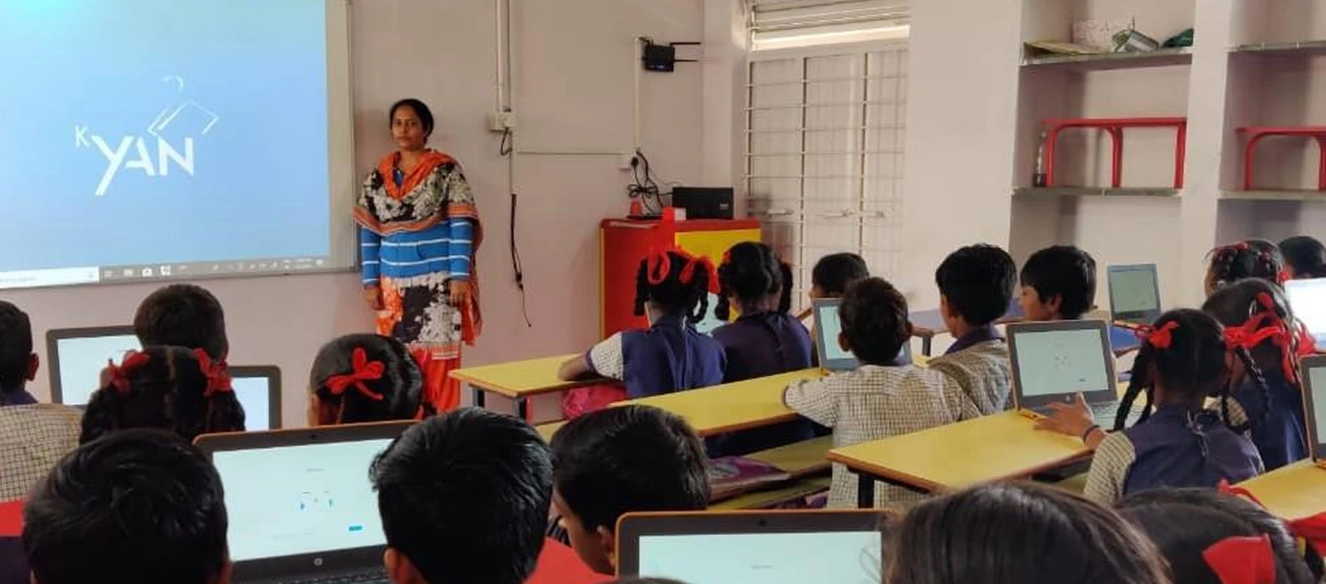 Teacher standing by a projector screen while students in uniform work on laptops in a classroom