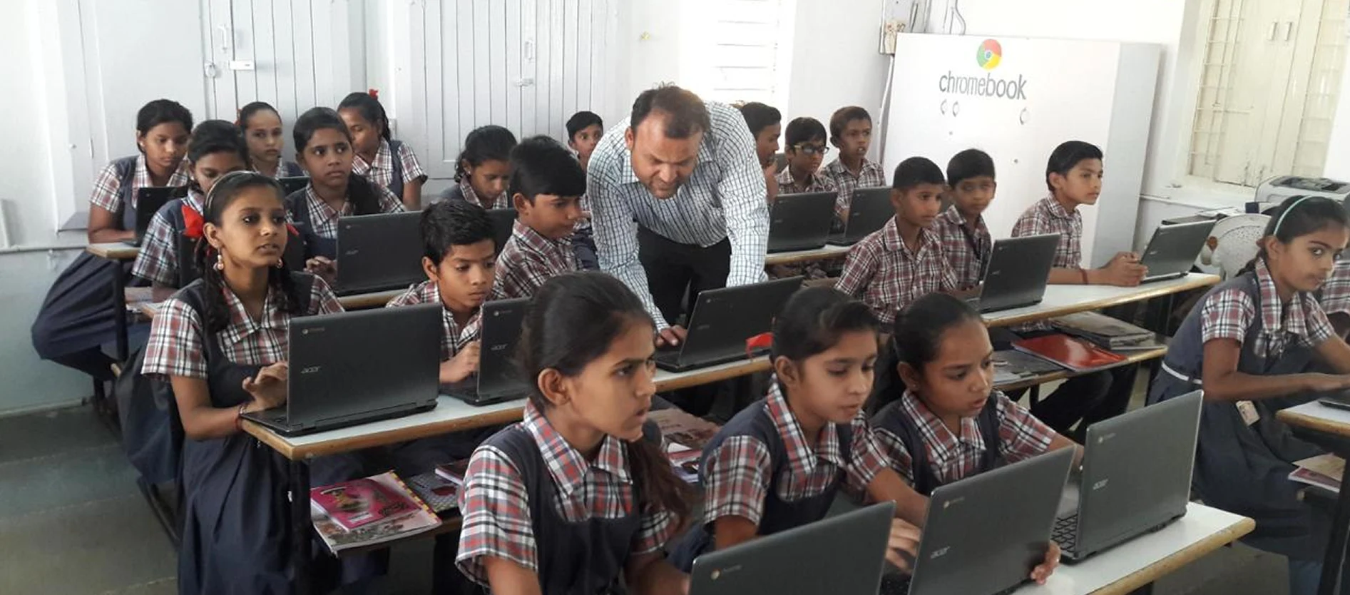 Teacher helping students in school uniform working on Chromebooks in a classroom