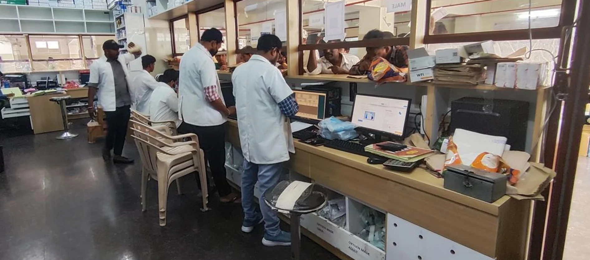 Pharmacy staff in white coats working at computers behind a busy hospital dispensing counter