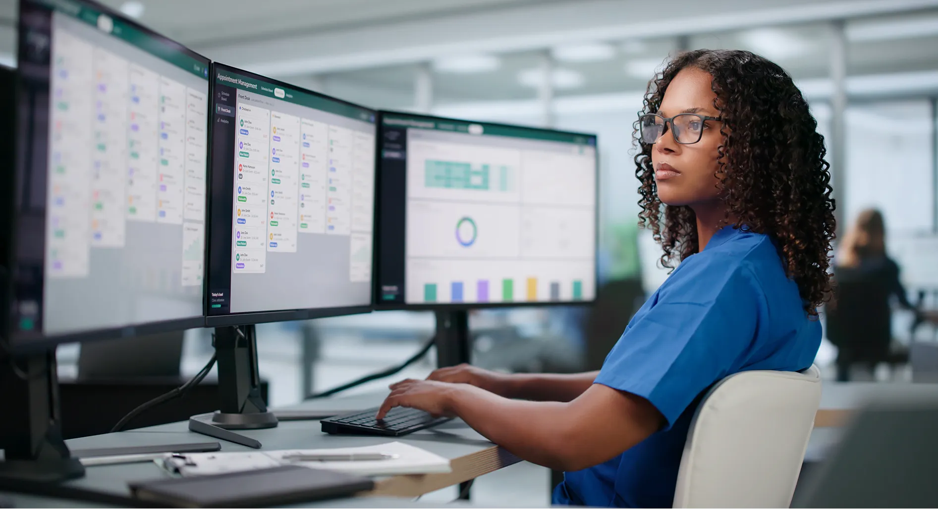 Hospital staff member working at a desk with three monitors showing the kNODE appointment dashboard