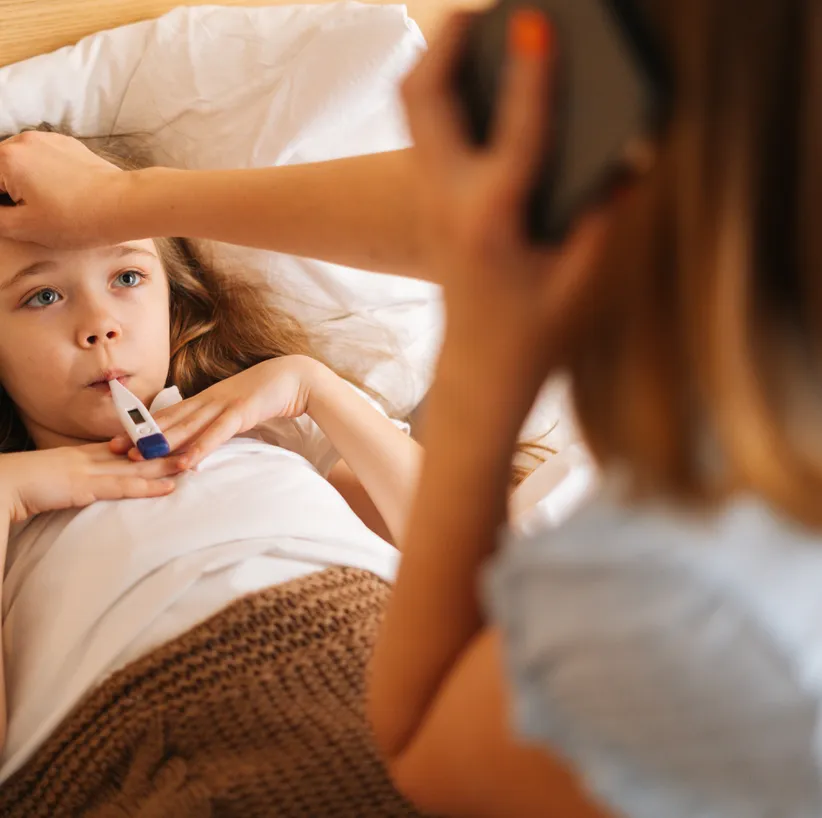 Mother checking the temperature of her daughter lying in bed while on the phone