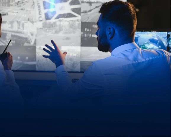 Two security professionals discussing surveillance feeds on large screens in a control room