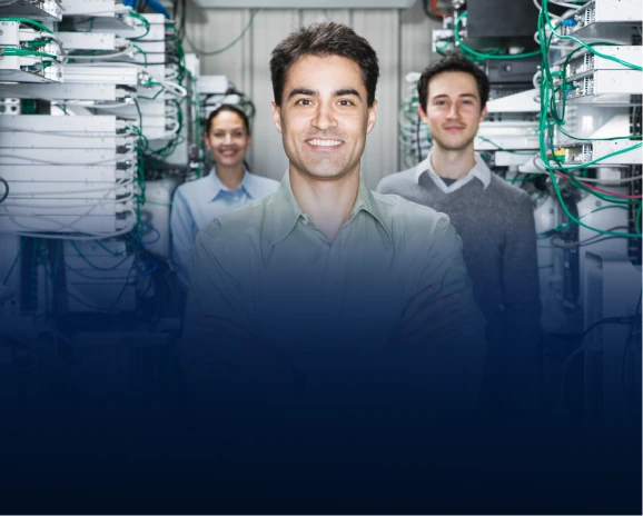 Three IT professionals smiling in a server room with racks of networking equipment